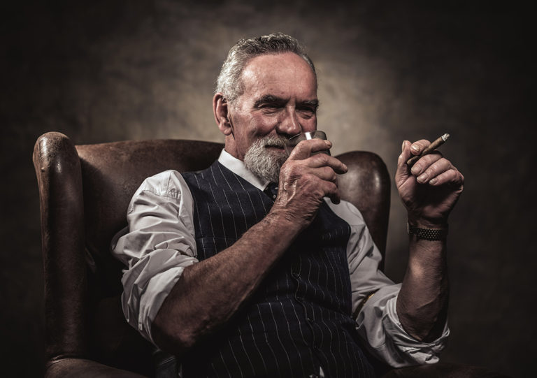 In chair sitting senior business man with cigar and whisky. Gray hair and beard wearing blue striped gilet and tie. Against brown wall.